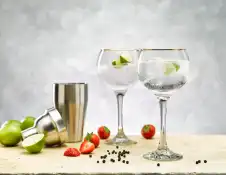 Two gin and tonics in goblets, a cocktail shaker with limes, strawberries and pepp ercornson the counter 
